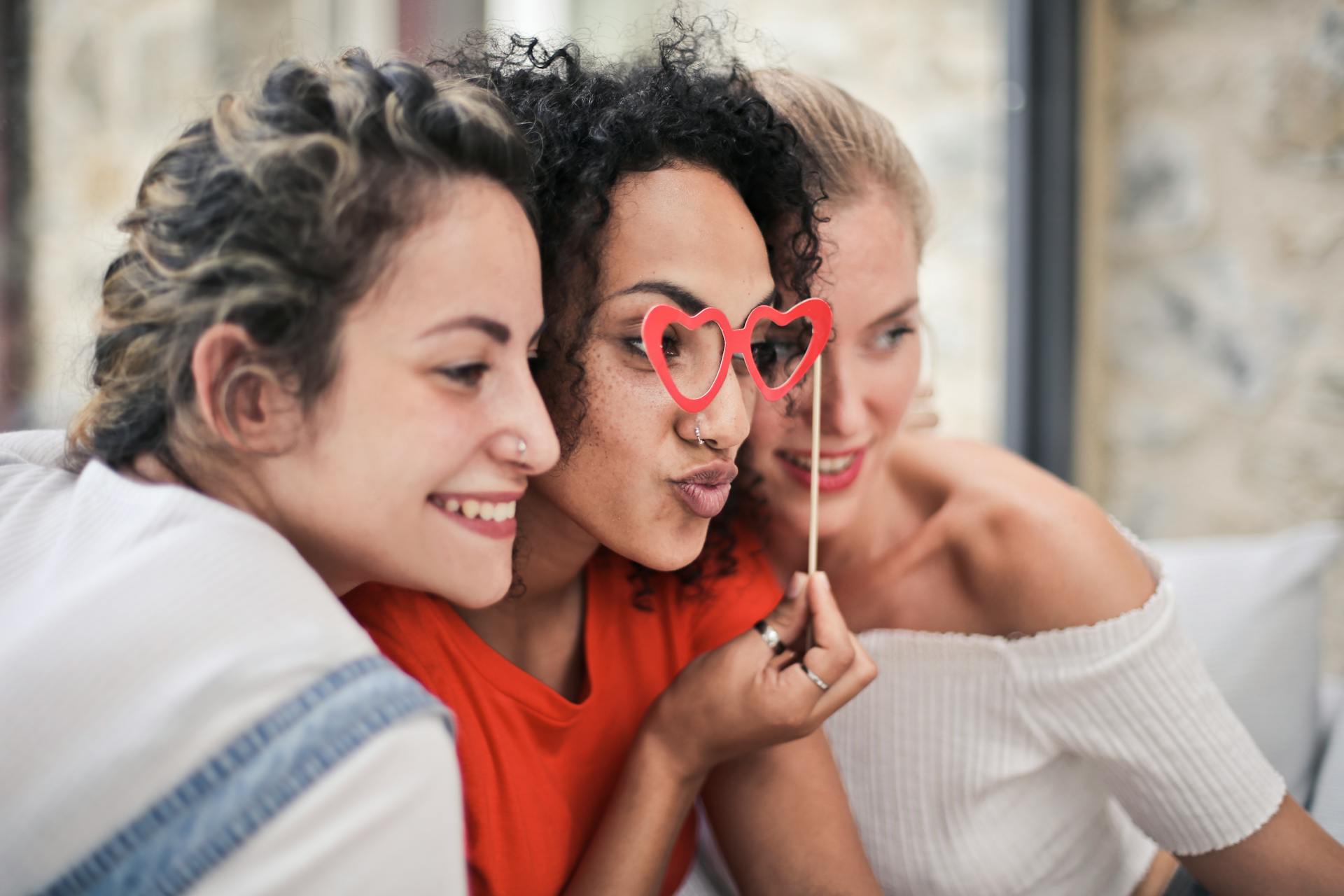 Three smiling friends celebrating Galentine’s Day, with one holding heart-shaped glasses.